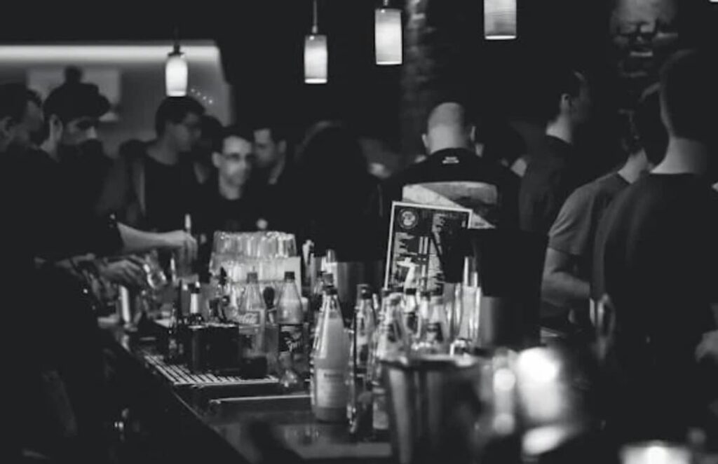 A blurry black and white photo of people at a bar with drinks and bottles in the foreground